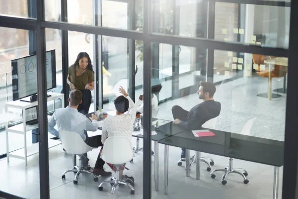 Office meeting with business professionals watching a presentation on a screen, engaging with questions, opinions, and curiosity during a workshop. In a glass-walled boardroom, a speaker leads a team discussion focused on corporate updates, review, and training information.